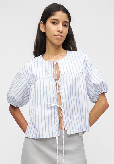Γυναίκα που φοράει μπλούζα με λευκές και μπλε κάθετες ρίγες, φουσκωτά μανίκια και τρεις μπροστινές δεσμεύσεις, συνδυασμένη με ανοιχτό γκρι παντελόνι.