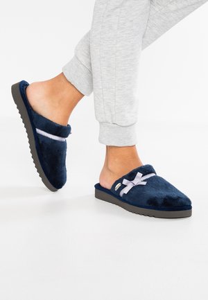 Marineblauwe fuzzy slippers met een zachte textuur, voorzien van een lavendelkleurige lintaccent en een platte, gestructureerde zool. Het ontwerp is open aan de achterkant.