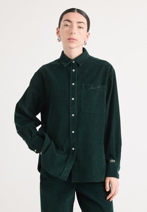 Camicia in velluto a coste verde scuro con colletto a punta, maniche lunghe, chiusura con bottoni, taschino sul petto e logo ricamato sulla tasca.