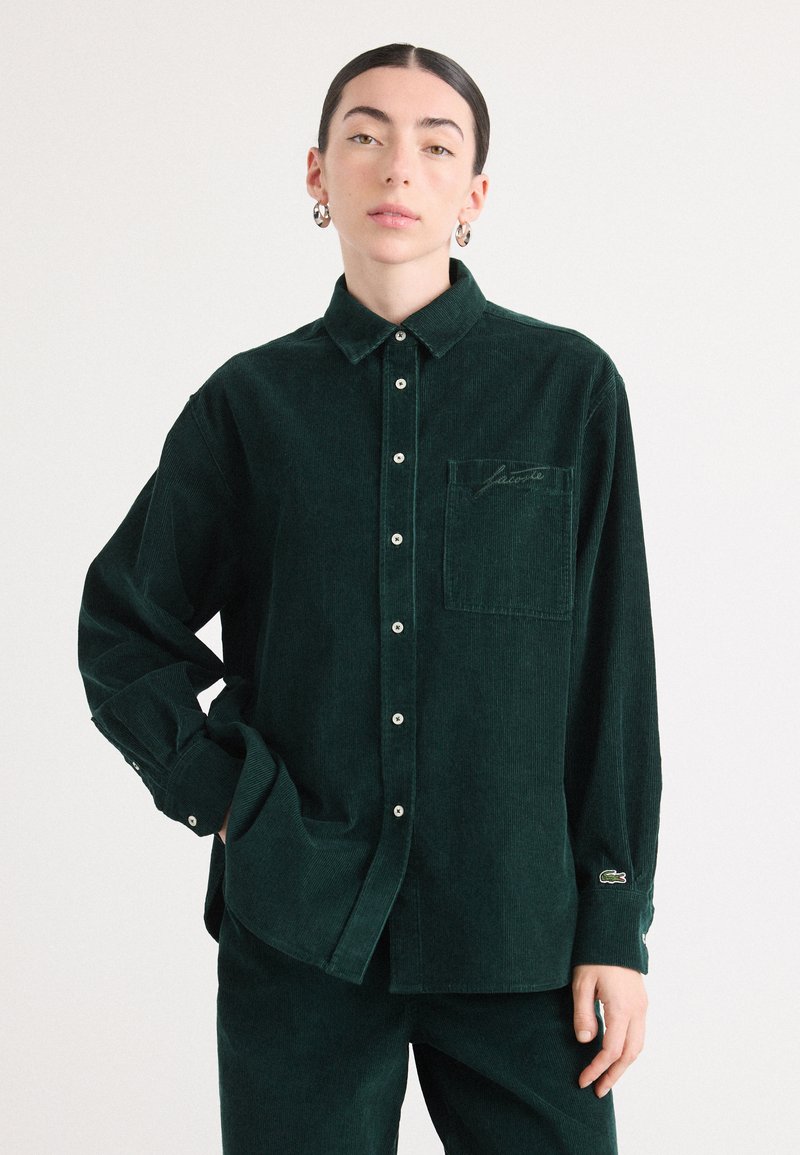Camicia in velluto a coste verde scuro con colletto a punta, maniche lunghe, chiusura con bottoni, taschino sul petto e logo ricamato sulla tasca.