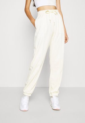 Femme portant un pantalon jogger crème taille haute avec chevilles élastiques et baskets blanches, debout avec une main dans la poche.