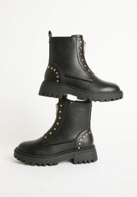Botines de cuero negro, con suela gruesa, adornos de estrellas doradas y remaches, y cremallera lateral, exhibidos sobre un fondo blanco liso.