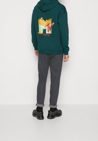 Sweat à capuche vert avec un graphisme de frites et le logo "MTV" au dos. Associé à un pantalon gris à fines rayures et des baskets noires. Coupe décontractée dans l'ensemble.