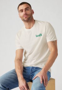 T-shirt en coton crème avec un logo Levi's vert sur la poitrine gauche, associé à un jean en denim bleu clair.