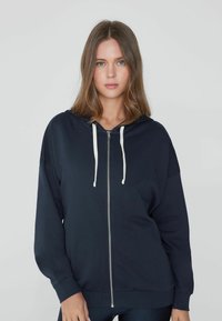 Marineblaue Kapuzenjacke mit Reißverschluss, ausgestattet mit weißen Kordeln, überschnittenen Schultern und gerippten Bündchen. Weicher Stoff mit glatter Textur.