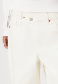 Jeans in denim bianco con un classico design a cinque tasche, tessuto testurizzato, dettagli in rame e chiusura con bottone metallico.