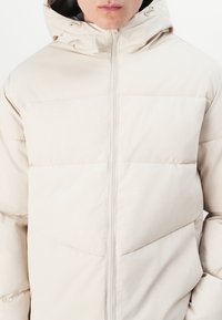 Chaqueta acolchada beige con capucha, presenta costuras horizontales, cremallera frontal y bolsillos laterales. Textura suave y diseño ligero.