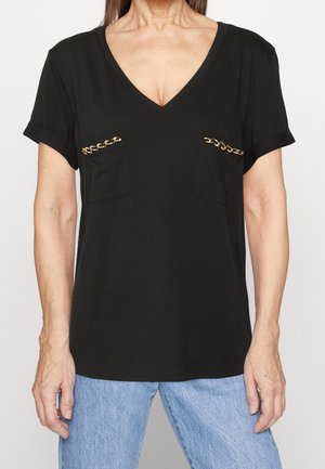 Femme portant un haut noir à col en V manches courtes avec des détails de chaîne dorée sur les poches de poitrine et un jean bleu clair.
