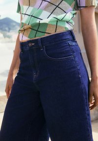 Jeans gelap berwarna biru dengan pinggang tinggi dan desain kaki lurus, menampilkan tombol yang terlihat dan detail jahitan. Dipadukan dengan atasan lengan pendek bermotif.