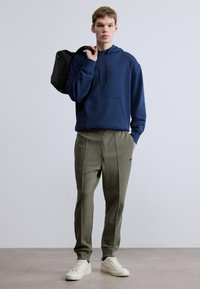 Tengerészkék kapucnis pulóver elülső zsebbel, olíva zöld szűkített nadrággal és fehér tornacipővel párosítva. A modell fekete táskát visz.