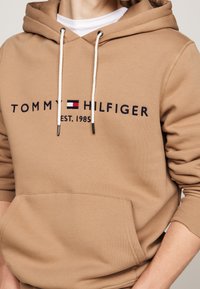 Felpa con cappuccio beige con tasca a marsupio frontale, cappuccio con coulisse e logo ricamato "TOMMY HILFIGER EST. 1985" in blu scuro e rosso.