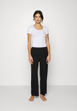 T-shirt ajusté blanc à manches courtes associé à un pantalon large noir, sur un fond blanc uni. Le modèle est pieds nus.