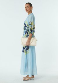 Hellblaues, blühendes Maxikleid mit Puffärmeln, das Navy- und Grüntöne aufweist. Das Modell hält eine cremefarbene Handtasche und trägt weiße Sandalen mit Riemchen.