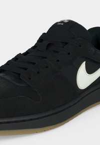 Baskets basses Nike SB noires avec logo swoosh blanc, lacets noirs et semelle couleur gomme, inclinées pour montrer les détails latéraux et avant.