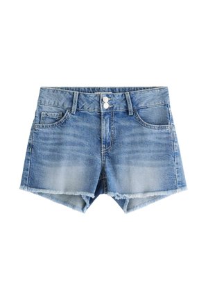Teenagepige med langt krøllet hår iført hvid skulderbluse, denimshorts og pinke sneakers, stående med den ene hånd i lommen.