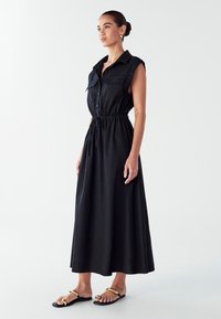 Schwarzes, ärmelloses Maxikleid mit durchgehender Knopfleiste, zwei Brusttaschen, verstellbarem Taillengurt und fließendem Saum; kombiniert mit flachen Sandalen.