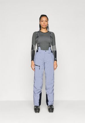 Pantalons de neige lilas clair avec bretelles noires réglables, poche latérale avec fermeture éclair, et poignets renforcés. Associé à un haut ajusté à manches longues noir.