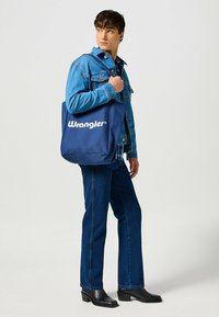 Un jeune homme portant une veste en jean et un jean tient un sac fourre-tout bleu Wrangler sur son épaule, regardant sur le côté.