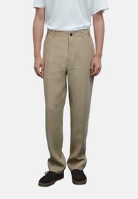 Pantaloni in lino beige con taglio dritto, dotati di chiusura frontale con bottone, passanti per cintura e tasche laterali. Indossati con una semplice maglietta bianca.