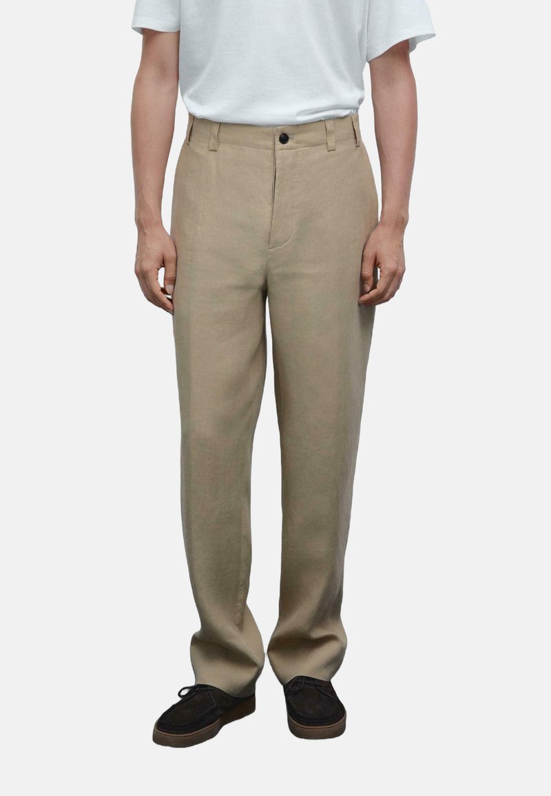 Pantaloni in lino beige con taglio dritto, dotati di chiusura frontale con bottone, passanti per cintura e tasche laterali. Indossati con una semplice maglietta bianca.
