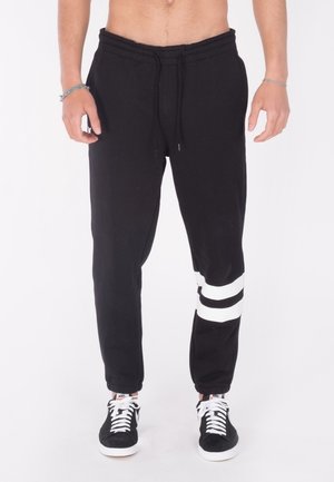 Mężczyzna w czarnych spodniach dresowych z dwoma białymi paskami na prawej nodze oraz czarnych butach Nike z białymi sznurowadłami, stojący na białym tle.