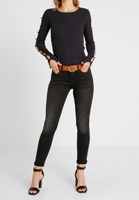 Femme portant un haut noir à manches longues côtelé avec des découpes à manches boutonnées, un jean skinny noir avec une ceinture marron et des sandales à talons noirs.