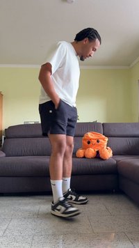 Homme en chemise blanche et shorts noirs se tenant à l'intérieur près d'un canapé gris foncé avec un octopus en peluche orange dessus.