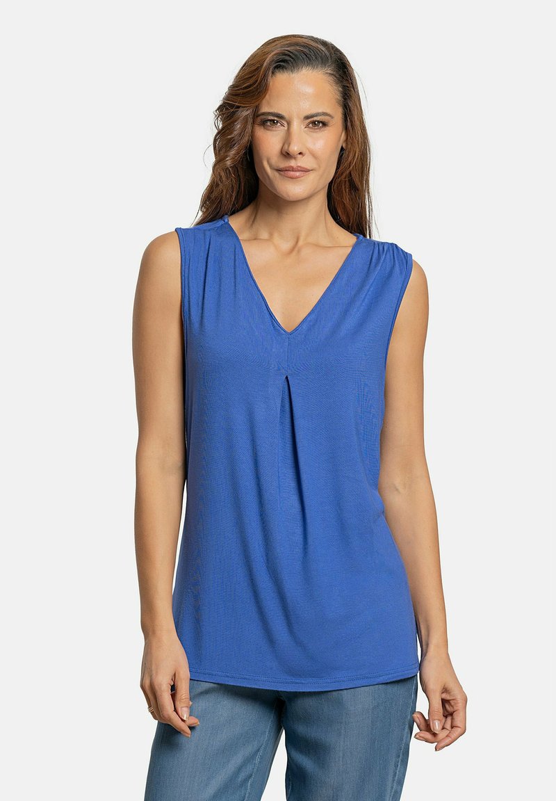 Top sin mangas azul con escote en V, detalle de pliegues en la parte delantera y tejido suave. Combinado con pantalones de mezclilla claros. Textura suave.