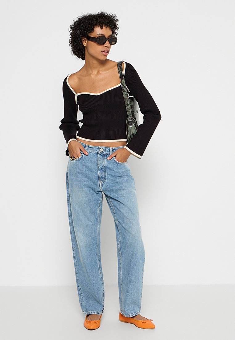 Schwarzes, geripptes Langarmoberteil mit Rundhalsausschnitt und kontrastierenden weißen Details, kombiniert mit hellblauen Weitbeinjeans und orangefarbenen Ballerinas.