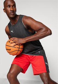 Svart mesh basketball tank top med hvite detaljer, kombinert med røde shorts med svarte sidestykker. Holder en oransje basketball.