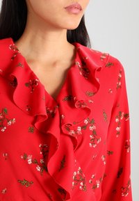Blusa rossa con scollatura arricciata, decorata con una stampa floreale in rosa e verde. Realizzata in tessuto morbido, il design mette in risalto colori vivaci e dettagli.