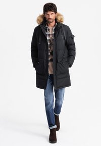 Cappotto puff al nero con cappuccio foderato in pelliccia, due tasche frontali e cerniere. Indossato sopra una camicia a quadri, abbinato a jeans blu e stivali marroni.