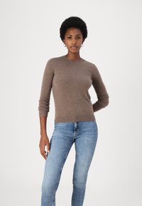 Maglione lavorato a maglia marrone con scollatura tonda, maniche lunghe e polsini a coste, abbinato a jeans blu attillati. Texture liscia.
