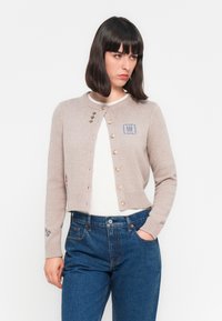 Lichtbeige cropped cardigan met geborduurde details, ronde halslijn en knoopsluiting. Draagt over een witte blouse en gecombineerd met blauwe jeans.
