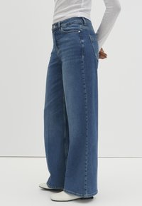 Jean en denim bleu à jambes larges avec taille haute, cinq poches et des coutures minimales. Le tissu a une finition lisse et légèrement texturée.