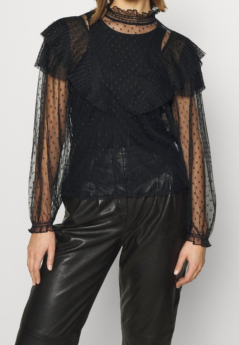 Schwarze transparente Bluse mit Polka-Dots-Muster, Rüschenelemente an den Schultern, hochgeschlossener Kragen mit Rüschensaum, kombiniert mit glänzenden schwarzen Lederhosen.
