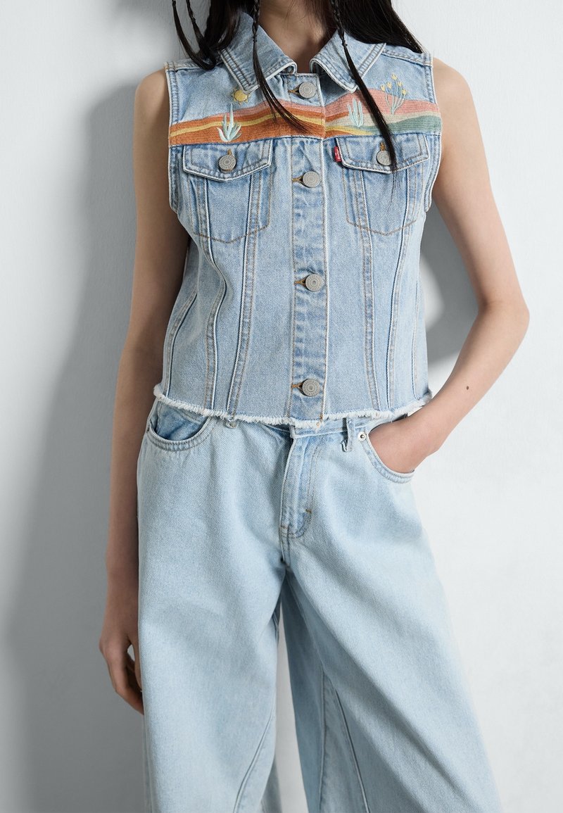 Bezrekawnikowa jasnoniebieska kamizelka dżinsowa z haftowanym pejzażem pustynnym, zestawiona z luźnymi jasnoniebieskimi dżinsami, modelka z jedną ręką w kieszeni.