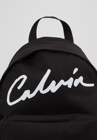 Czarny plecak z materiału, z dużym białym logo na froncie, o zaokrąglonym kształcie i uchwytem górnym do noszenia.