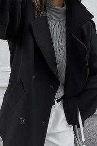 Cappotto di lana nera con ampio colletto e bottoni doppi, indossato sopra un maglione grigio a trecce, abbinato a pantaloni bianchi e una piccola borsa bianca.