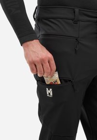 Czarne spodnie softshell z kieszenią na zamek, zaprojektowane do użytku na świeżym powietrzu, z widocznym logo i ręką wkładającą opakowanie przekąski.