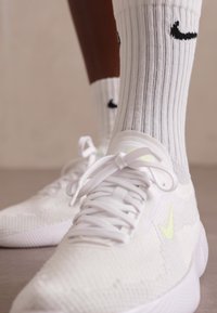 White athletic sneakers with textured knit upper, flat laces, and small neon yellow logo. Paired with white ribbed socks featuring black logo.