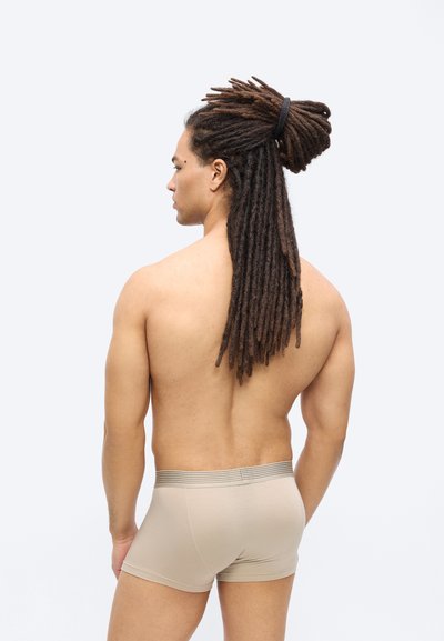 Άνδρας με μακριές κοτσίδες δεμένες προς τα πίσω, φαίνεται από την πλάτη, φοράει μπεζ ανδρικά εσώρουχα τύπου μποξέρ σε απλό ανοιχτό φόντο.