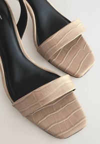 Talons en simili cuir crocodile beige avec un design à talon carré et deux larges sangles, associés à une semelle intérieure noire élégante.