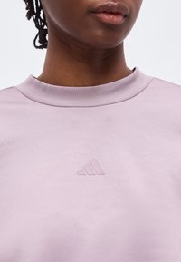 Κοντινό πλάνο ενός ατόμου που φοράει ένα ανοιχτό μοβ πουκάμισο με ένα μικρό ανάγλυφο λογότυπο της Adidas τοποθετημένο στο κέντρο κάτω από το λαιμόκοψη.