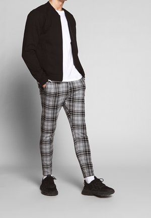 Veste bomber noire sur un t-shirt blanc, accompagnée de pantalons à carreaux gris et noirs. Des baskets noires complètent la tenue.