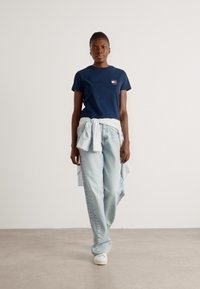Marinecrewneck t-shirt, lys blå løstsiddende jeans, med en stribet skjorte knyttet om taljen; modellen bærer hvide sneakers.