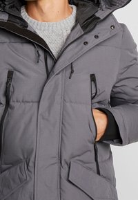 Giacca isolante grigia con cappuccio, dotata di zip frontale, accenti neri, multiple tasche e una superficie in tessuto testurizzato.