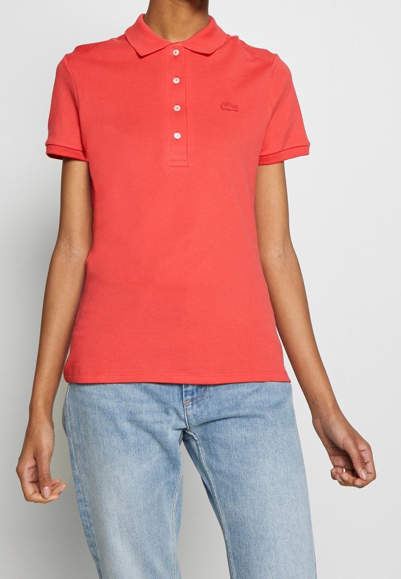 Personne portant un polo rouge corail à manches courtes et un jean bleu clair sur un fond uni.