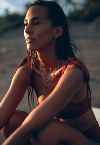 Rote Badebekleidung mit einem geknoteten Schulterdesign, geschichteten silbernen Halsketten mit verschiedenen Anhängern und nassen, strukturierten Haaren.