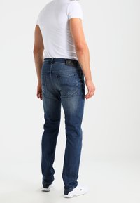 Vaqueros de mezclilla azul oscuro con corte ajustado, diseño de cinco bolsillos y una etiqueta de logo en la parte posterior. Llevados con una camiseta blanca ajustada y zapatillas blancas.